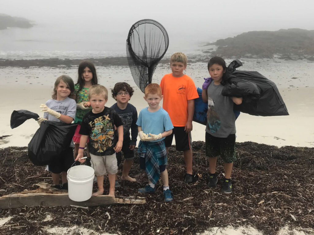 Children stand in a coastal landscape on an overcast day. Some of the students hold objects, including a net, black plastic trash bags, and a bucket.
