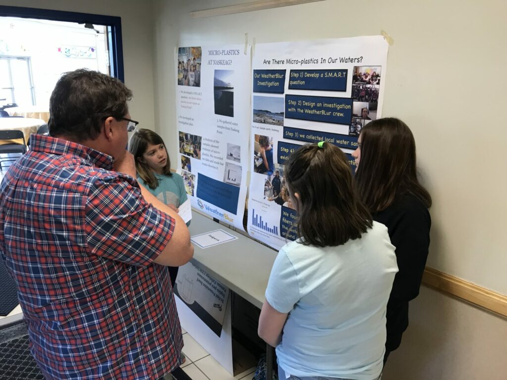 An adult looks at a poster presentation titled "Are There Micro-plastics in Our Waters?" as students describe the two poster, which includes charts and images.